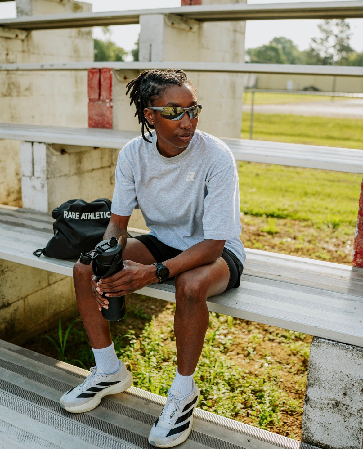 Athlete sitting on a metal bench holding a water bottle, wearing Rare Athletics tee,  sunglasses, and athletic shoes.